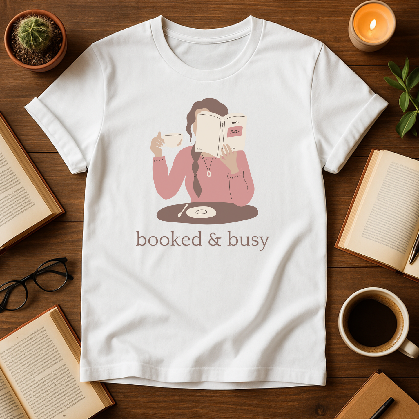White t-shirt with a graphic of a person reading a book and holding a coffee cup, with the text 'booked & busy', on a wooden surface with books and a candle.
